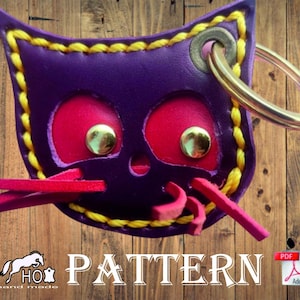 Pode incluir: Chaveiro de couro roxo em forma de gato com detalhes em feltro vermelho, costuras amarelas e um anel de metal dourado. O chaveiro tem a palavra "PATTERN" escrita em letras grandes abaixo. A imagem também inclui um ícone PDF.