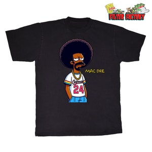 May include: Black t-shirt featuring a cartoon character with a large afro, gold chain, and the text "MAC DRE". The character wears a white, rainbow-striped jersey with "California 24" on the front. The shirt has a crew neck.