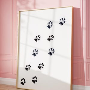 May include: A framed print with a white background and black paw prints in a scattered pattern. The frame is gold.