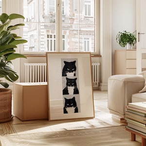 May include: A framed art print featuring three black and white cat portraits. Each cat has a different expression, with the top cat appearing angry. The artwork is displayed in a light-colored room with a large window and a potted plant.
