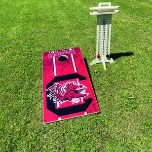 Puede incluir: Un juego de cornhole rojo y negro con el logotipo de los Gamecocks de la Universidad de Carolina del Sur. El tablero de juego tiene una "C" negra con un gallo rojo dentro. El tablero de puntuaci&oacute;n es blanco con n&uacute;meros negros.