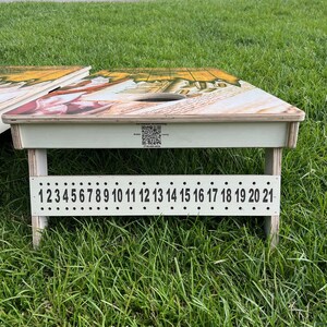 Puede incluir: Un juego de cornhole de madera blanca con un dise&ntilde;o colorido en la parte superior. El juego tiene un sistema de puntuaci&oacute;n numerado del 1 al 21.