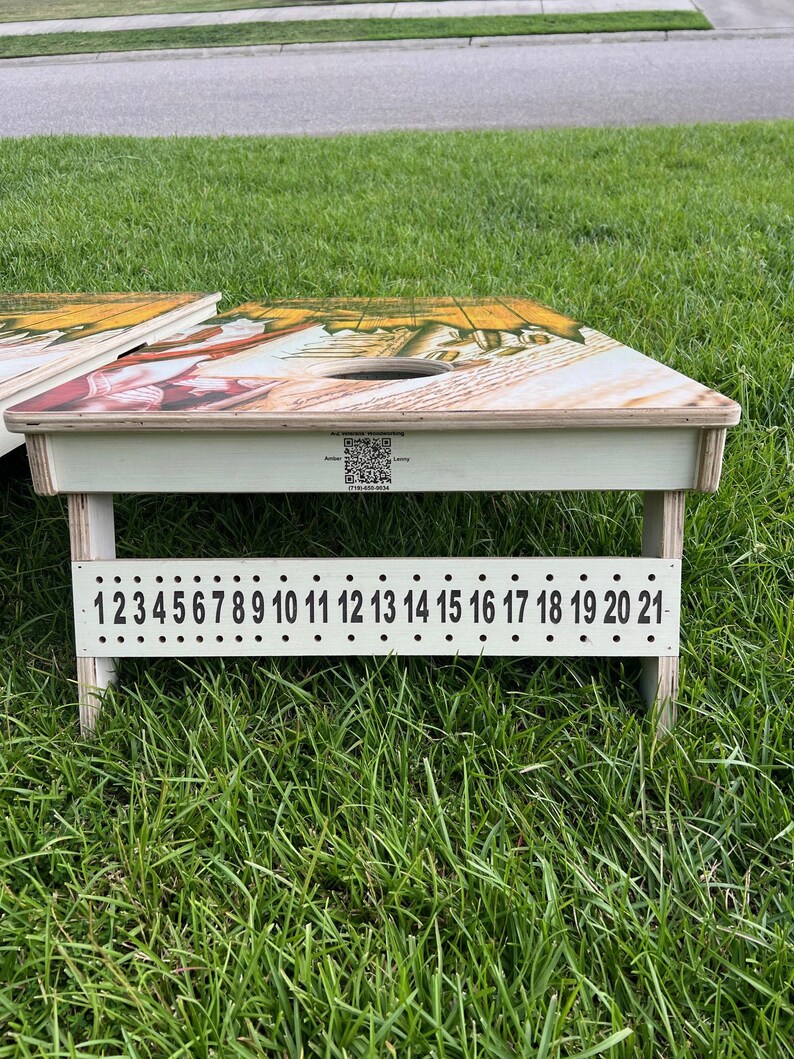 Puede incluir: Un juego de cornhole de madera blanca con un dise&ntilde;o colorido en la parte superior. El juego tiene un sistema de puntuaci&oacute;n numerado del 1 al 21.