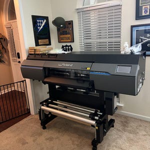 Puede incluir: Una impresora de gran formato Roland TrueVIS VG2-640 negra y gris con una pantalla digital. La impresora est&aacute; en un soporte con dos rodillos y se encuentra en una habitaci&oacute;n con suelo de moqueta.