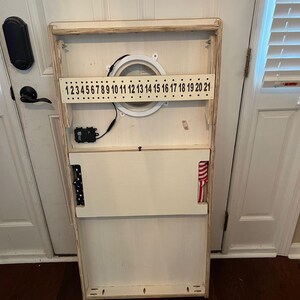 Puede incluir: Un tablero de juego de madera blanco con una abertura circular y un sistema de puntuaci&oacute;n numerado. El tablero tiene una tela a rayas roja, blanca y azul unida al lado.