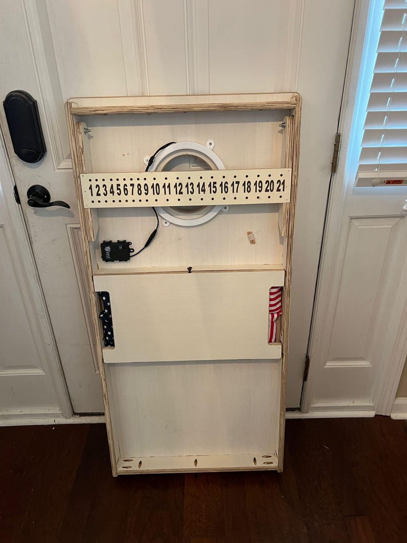 Puede incluir: Un tablero de juego de madera blanco con una abertura circular y un sistema de puntuaci&oacute;n numerado. El tablero tiene una tela a rayas roja, blanca y azul unida al lado.