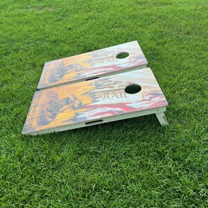 Puede incluir: Dos tablas de cornhole de madera con un dise&ntilde;o patri&oacute;tico. Las tablas est&aacute;n sobre una superficie de hierba verde. Las tablas tienen un dise&ntilde;o rojo, blanco y azul con las palabras "We the People" impresas en ellas.