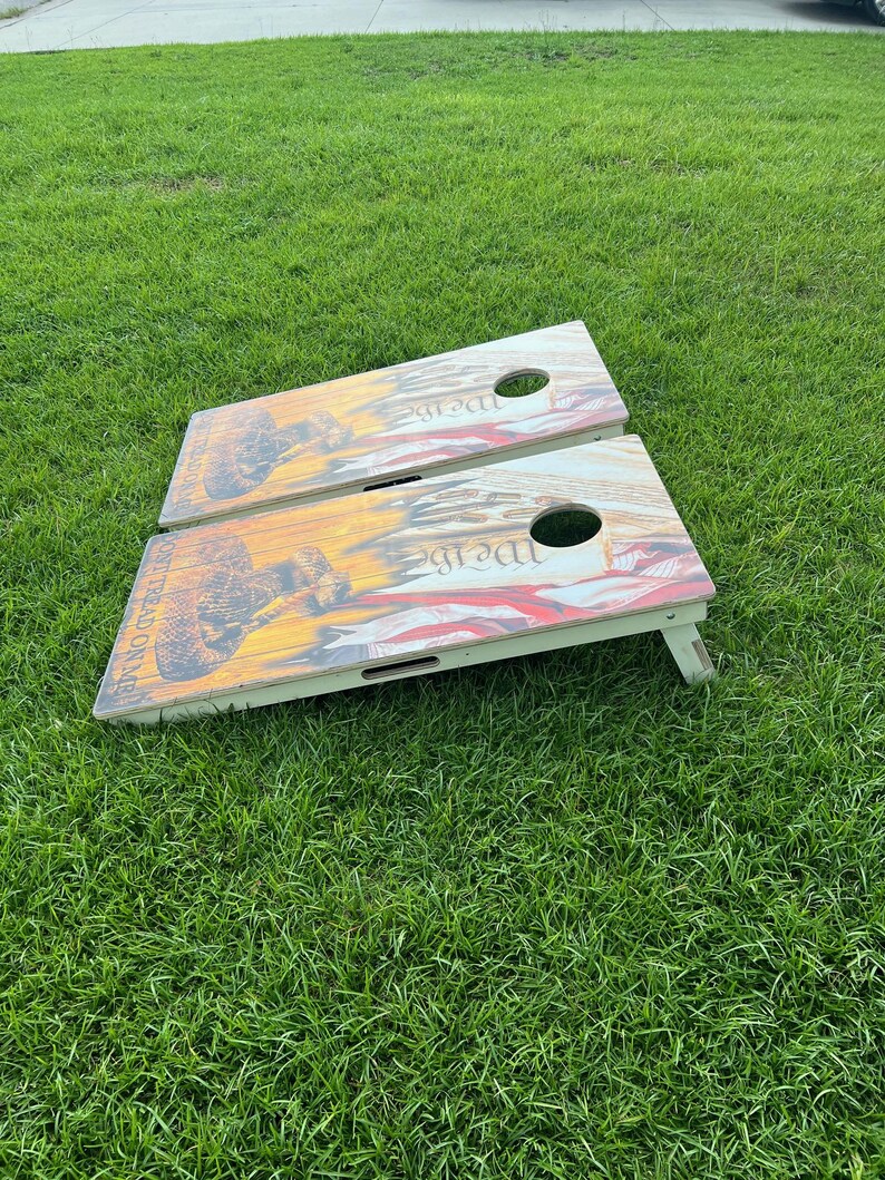 Puede incluir: Dos tablas de cornhole de madera con un dise&ntilde;o patri&oacute;tico. Las tablas est&aacute;n sobre una superficie de hierba verde. Las tablas tienen un dise&ntilde;o rojo, blanco y azul con las palabras "We the People" impresas en ellas.