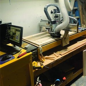 Puede incluir: Primer plano de una m&aacute;quina de fresado CNC con un panel de madera en la mesa. La m&aacute;quina est&aacute; conectada a un ordenador con un monitor que muestra un dise&ntilde;o. La fresadora est&aacute; en un taller con herramientas y restos de madera en el suelo.
