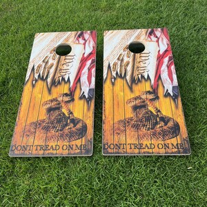 Puede incluir: Dos tablas de cornhole de madera con un dise&ntilde;o patri&oacute;tico. Las tablas presentan una serpiente de cascabel, una bandera estadounidense desgarrada y las palabras "Don't Tread On Me".