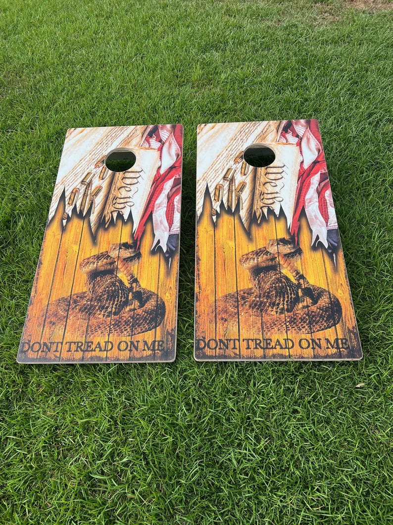 Puede incluir: Dos tablas de cornhole de madera con un dise&ntilde;o patri&oacute;tico. Las tablas presentan una serpiente de cascabel, una bandera estadounidense desgarrada y las palabras "Don't Tread On Me".
