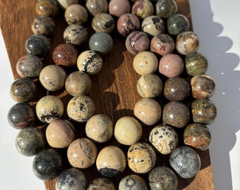 May include: A strand of polished gemstone beads in various earth tones, including beige, brown, and gray, displayed on a wooden board. The beads are round and have unique patterns and textures, suggesting natural stone. The beads are strung together with a thin, clear cord.
