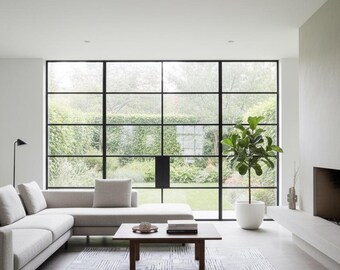 May include: A modern living room with a light gray sectional sofa, a square wooden coffee table, and a patterned rug in shades of gray and blue. A large window with black frames overlooks a green outdoor space. A potted plant adds a touch of nature.