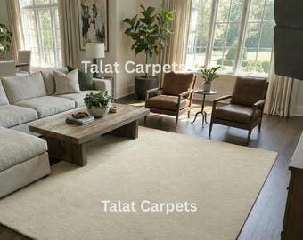 May include: A neutral-toned area rug in a living room setting. The rug is rectangular and covers a large portion of the floor. The room features a sectional sofa, two leather armchairs, and a wooden coffee table. The text "Talat Carpets" is visible.