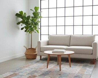 Op de afbeelding: Een moderne woonkameropstelling met een beige bank, een ronde houten salontafel en een patroonkleed. Een groot raam en een potplant dragen bij aan de esthetiek van de kamer. Het kleed heeft een geometrisch ontwerp met blauwe, bruine en witte strepen.