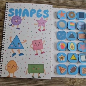 Può includere: Un libro educativo rilegato a spirale intitolato "SHAPES" con illustrazioni colorate di varie forme, tra cui un cerchio, una stella, un triangolo e un quadrato, ognuno con un volto sorridente e arti. Ulteriori forme sono visualizzate su quadrati blu.