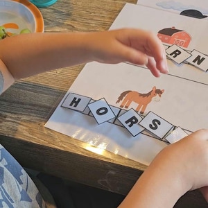 May include: Educational activity sheet with a brown horse illustration and letter tiles spelling "HORSES". A red barn illustration is also visible. The activity is on a white sheet of paper, with a child's hand interacting with the letters.