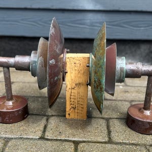 Puede incluir: Una escultura r&uacute;stica de estilo industrial con un bloque de madera central y componentes met&aacute;licos. Los elementos met&aacute;licos incluyen hojas circulares en forma de abanico y soportes cil&iacute;ndricos.