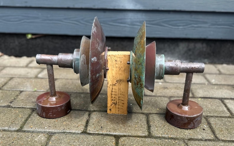 Puede incluir: Una escultura r&uacute;stica de estilo industrial con un bloque de madera central y componentes met&aacute;licos. Los elementos met&aacute;licos incluyen hojas circulares en forma de abanico y soportes cil&iacute;ndricos.