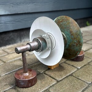 Puede incluir: Un objeto met&aacute;lico de estilo industrial con una base cil&iacute;ndrica y un brazo de soporte vertical. El objeto presenta una serie de discos blancos circulares y un disco m&aacute;s grande de color cobre envejecido. La pieza est&aacute; sobre una superficie de ladrillo.