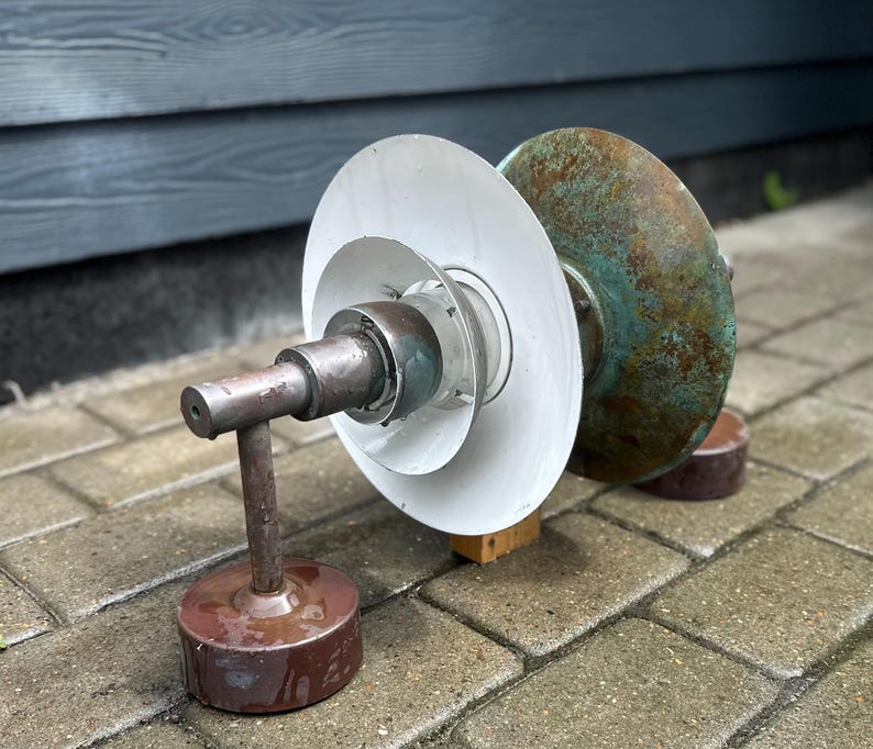 Puede incluir: Un objeto met&aacute;lico de estilo industrial con una base cil&iacute;ndrica y un brazo de soporte vertical. El objeto presenta una serie de discos blancos circulares y un disco m&aacute;s grande de color cobre envejecido. La pieza est&aacute; sobre una superficie de ladrillo.