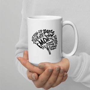 Puede incluir: Taza de cerámica blanca con un diseño negro de la palabra "EMDR" rodeada de otras palabras relacionadas con la terapia, como "bottom-up", "parts work", "somatic", "therapeutic", "relationship", "IFS", "connection", "emotional" y "neglect".