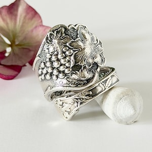 May include: A silver spoon ring featuring a detailed embossed design of a cluster of grapes and grape leaves on the wide top section, with a slender, patterned handle wrapping around to form the band of the ring.