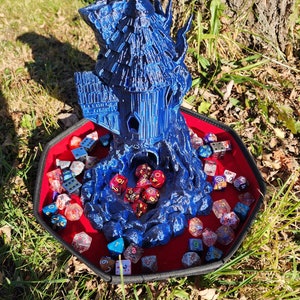 May include: A blue 3D printed tower with a thatched roof sits on a red felt dice tray with a variety of colorful dice. The tower is surrounded by a rocky base.