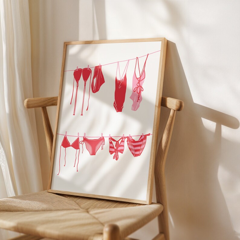 May include: A framed print featuring a minimalist illustration of various red and pink swimsuits hanging on a clothesline. The swimsuits include bikinis and one-piece suits, with a light beige background. The artwork is displayed in a light wood frame.