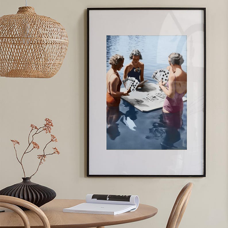 Puede incluir: Impresi&oacute;n art&iacute;stica enmarcada que muestra a tres personas con trajes de ba&ntilde;o vintage jugando a las cartas en el agua. La obra de arte est&aacute; enmarcada en un marco negro y se exhibe en una pared de color neutro. Una l&aacute;mpara colgante tejida cuelga sobre una mesa de madera.