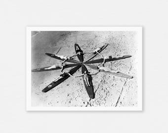 May include: Black and white photograph of people lying on surfboards arranged in a circular pattern on a sandy beach. The surfboards are radiating outwards from a central point, creating a star-like design.