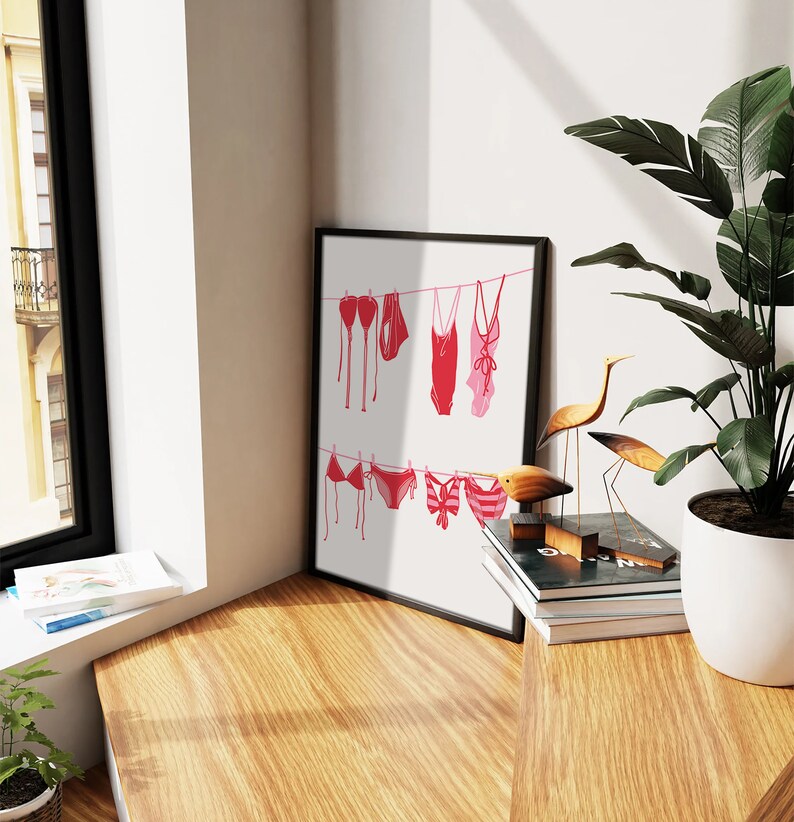 May include: A framed art print featuring a minimalist illustration of red and pink swimwear hanging on a clothesline. The artwork is displayed on a white background, with a black frame. The scene is set in a bright, well-lit room.