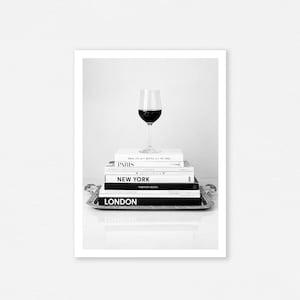 May include: A wine glass filled with red wine sits atop a stack of books on a silver tray. The books are labeled "Paris", "New York", and "London Fashion Book". The image is in black and white.
