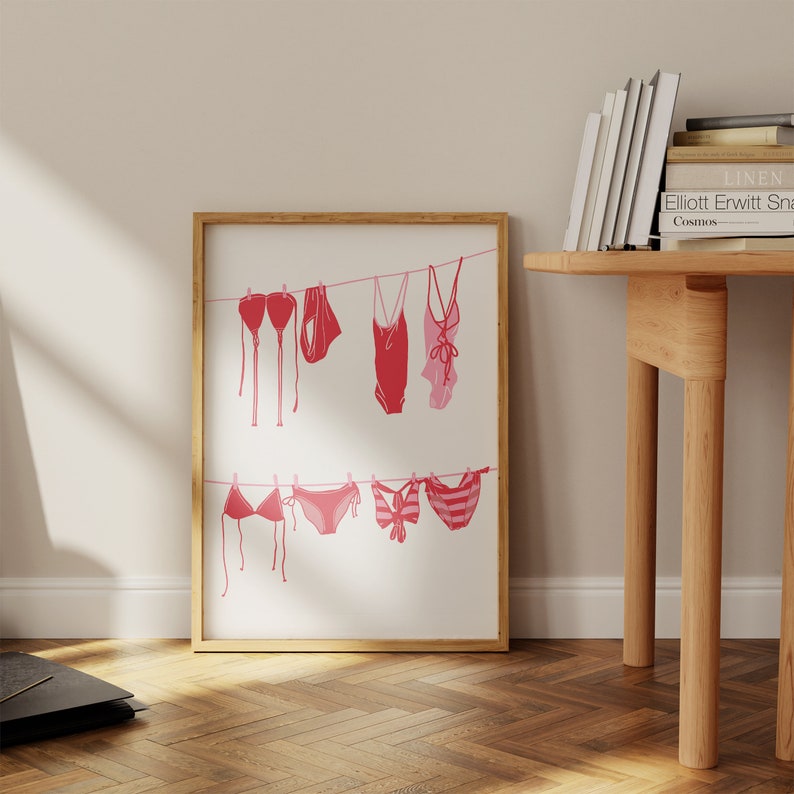 May include: A framed print featuring a line of pink and red swimsuits hanging on a clothesline. The swimsuits include bikinis and one-piece suits, with varying designs. The artwork is displayed in a room with a wooden floor and a wooden table.