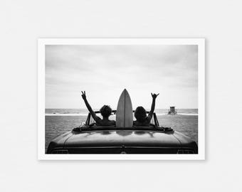 May include: Black and white photograph of a convertible car with two people and a surfboard. The people are in the front seats with their arms raised in the air. The surfboard is mounted on the car. The ocean and a lifeguard tower are in the background.