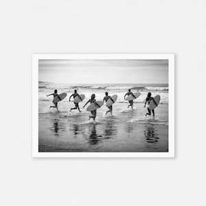 Op de afbeelding: Zwart-witfoto van zeven mensen die met surfplanken de oceaan in rennen. De afbeelding legt de beweging van de figuren vast terwijl ze naar de golven gaan, met water dat rond hun voeten spat. De scène speelt zich af op een strand onder een bewolkte hemel.
