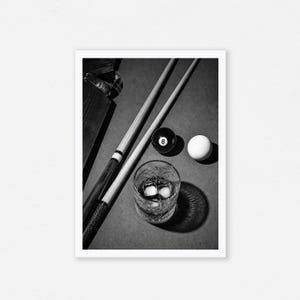 May include: Black and white photograph of a billiards scene. Two pool cues rest on a green felt table next to an eight ball, a white cue ball, and a rocks glass with ice cubes. The scene is lit with dramatic lighting.