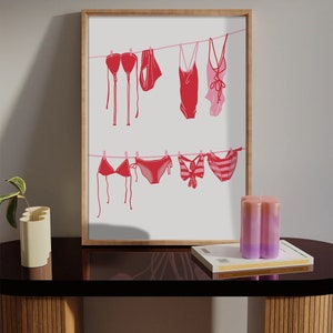 May include: A framed print featuring red and pink swimwear hanging on a clothesline. The artwork includes various bikini tops, bottoms, and one-piece swimsuits. The print is displayed on a dark table with a candle and a book.