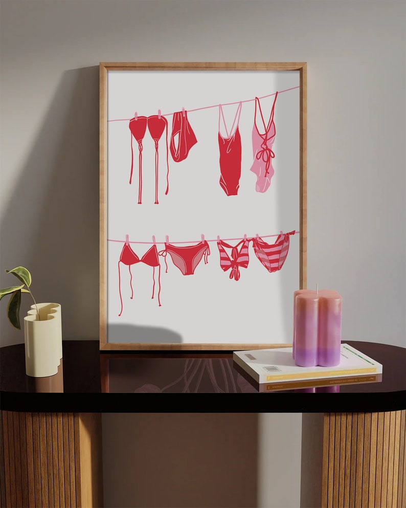 May include: A framed print featuring red and pink swimwear hanging on a clothesline. The artwork includes various bikini tops, bottoms, and one-piece swimsuits. The print is displayed on a dark table with a candle and a book.