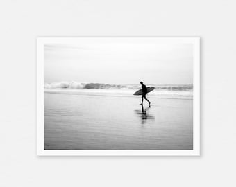 May include: Black and white photograph of a person walking on a beach carrying a surfboard. The ocean waves are visible in the background under a cloudy sky. The image is a minimalist depiction of a surfer.
