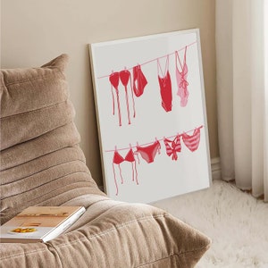 May include: A framed print featuring a minimalist illustration of various red and pink swimsuits hanging on clotheslines. The swimsuits include bikinis and one-piece suits, creating a summery and beach-themed aesthetic. The artwork is displayed against a neutral background.
