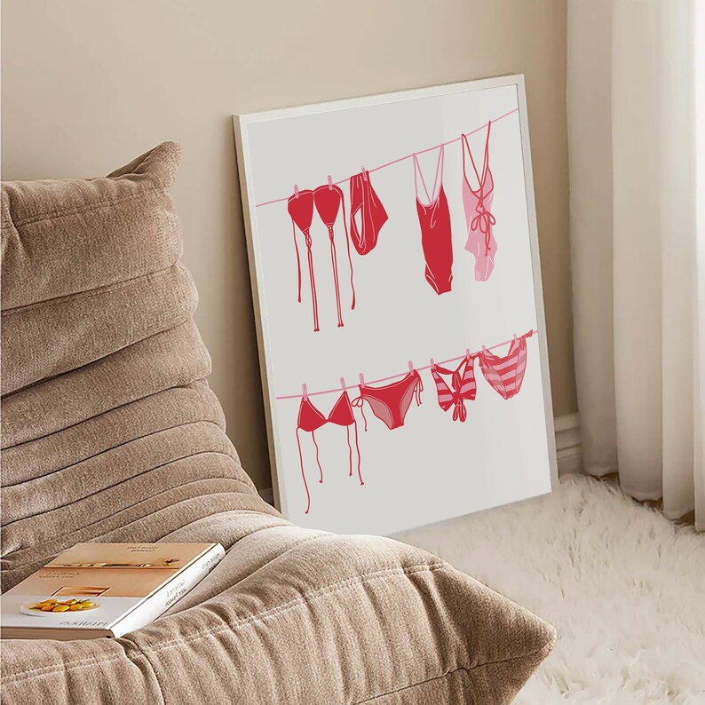 May include: A framed print featuring a minimalist illustration of various red and pink swimsuits hanging on clotheslines. The swimsuits include bikinis and one-piece suits, creating a summery and beach-themed aesthetic. The artwork is displayed against a neutral background.