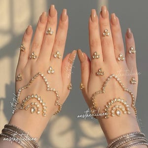 May include: Aesthetic hand jewelry set featuring gold-toned rings, hand chains, and bracelets. The rings and hand chains are adorned with clear crystals. The bracelets are silver-toned and stacked. The nails are painted a neutral color.