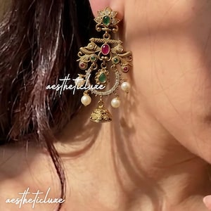 May include: A gold-toned chandelier earring featuring a floral top, intricate filigree metalwork, embedded red and green stones, clear crystals, and dangling white pearls with a small bell-shaped charm at the bottom, marked with the text aestheticluxe.