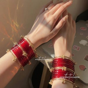 May include: A pair of red glass bangles with gold-colored accents and small dangling charms. The bangles are stacked on the wrist, with the text "aestheticluxe" visible on the image.