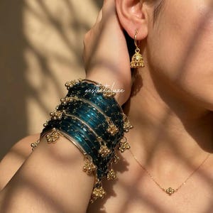 May include: A close-up of gold jewelry, including a bracelet, earrings, and a necklace. The bracelet is teal with gold accents and dangling gold charms. The earrings are small gold bells, and the necklace has a small gold pendant.