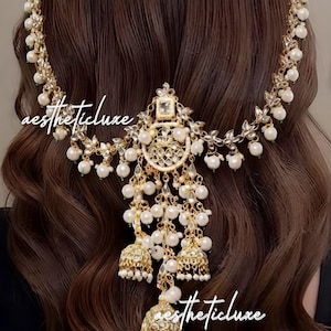 May include: A gold-toned hair accessory featuring a delicate chain adorned with white pearls and clear crystals. The centrepiece includes a circular pendant with dangling bell-shaped charms and additional pearl accents. The text "aestheticluxe" is visible.