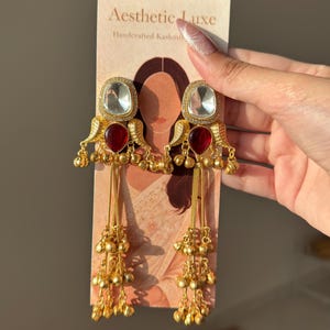 May include: Gold-toned chandelier earrings with clear and red gemstones. The earrings have a square, clear stone at the top, a teardrop-shaped red stone below, and dangling chains with small gold beads. The earrings are displayed on a card that reads "Aesthetic Luxe".