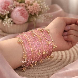 May include: A set of pink beaded bangles with gold-coloured details and hanging charms. The bangles are worn on a wrist, with a soft pink fabric in the background. A bouquet of pink flowers is also visible.