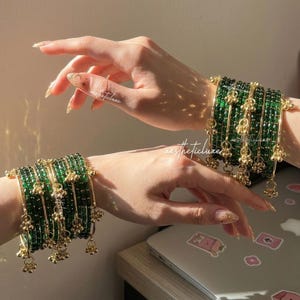 Könnte beinhalten: Zwei Hände zeigen mehrere grüne Glasarmreifen mit goldenen Akzenten und hängenden Charms. Die Armreifen sind gestapelt, wodurch ein geschichteter Effekt entsteht. Das Bild ist gut beleuchtet und hebt die Details des Schmucks hervor. Der Text "aestheticluxe" ist sichtbar.